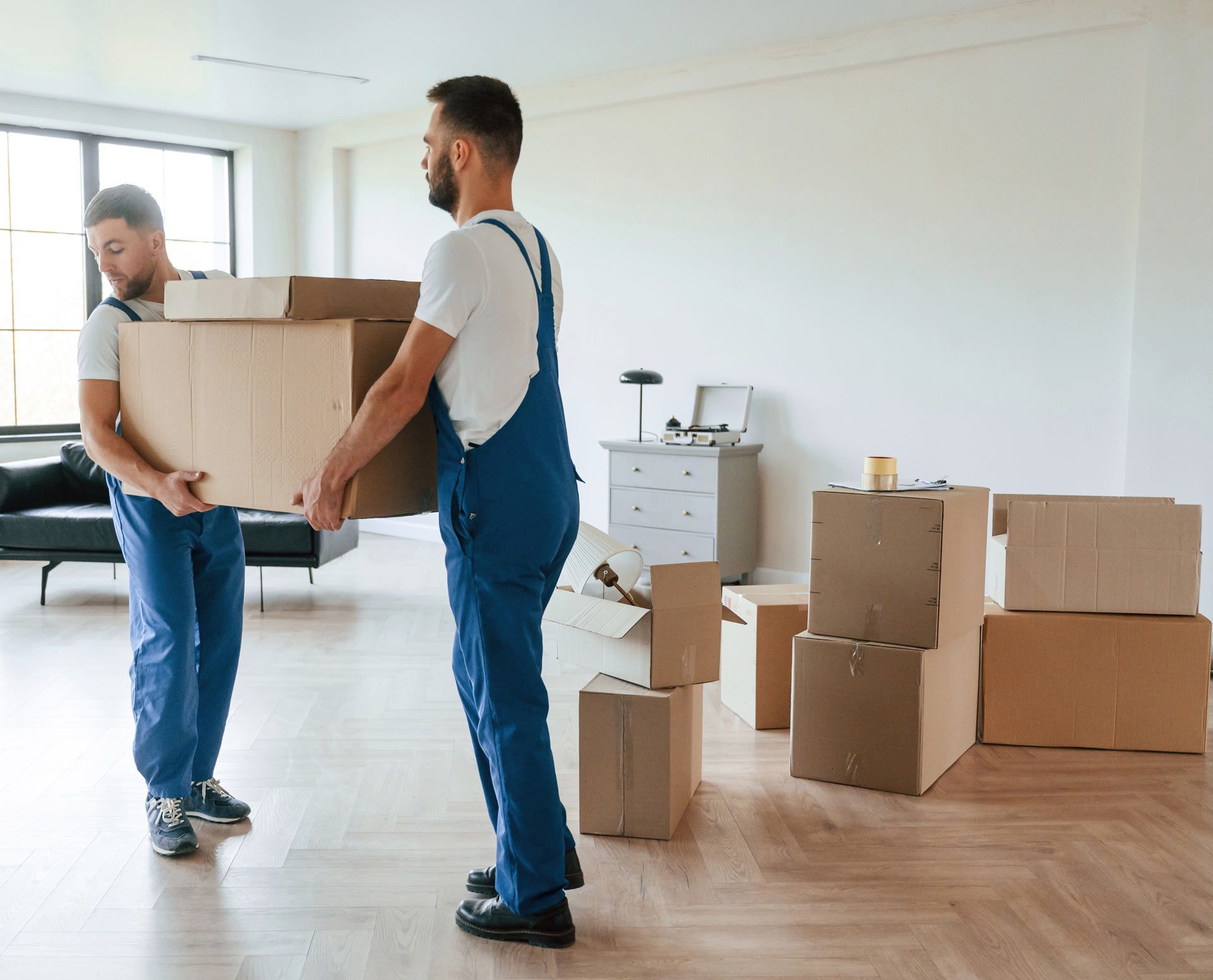 Two movers in blue coveralls carrying boxes in an empty room with other packed boxes.