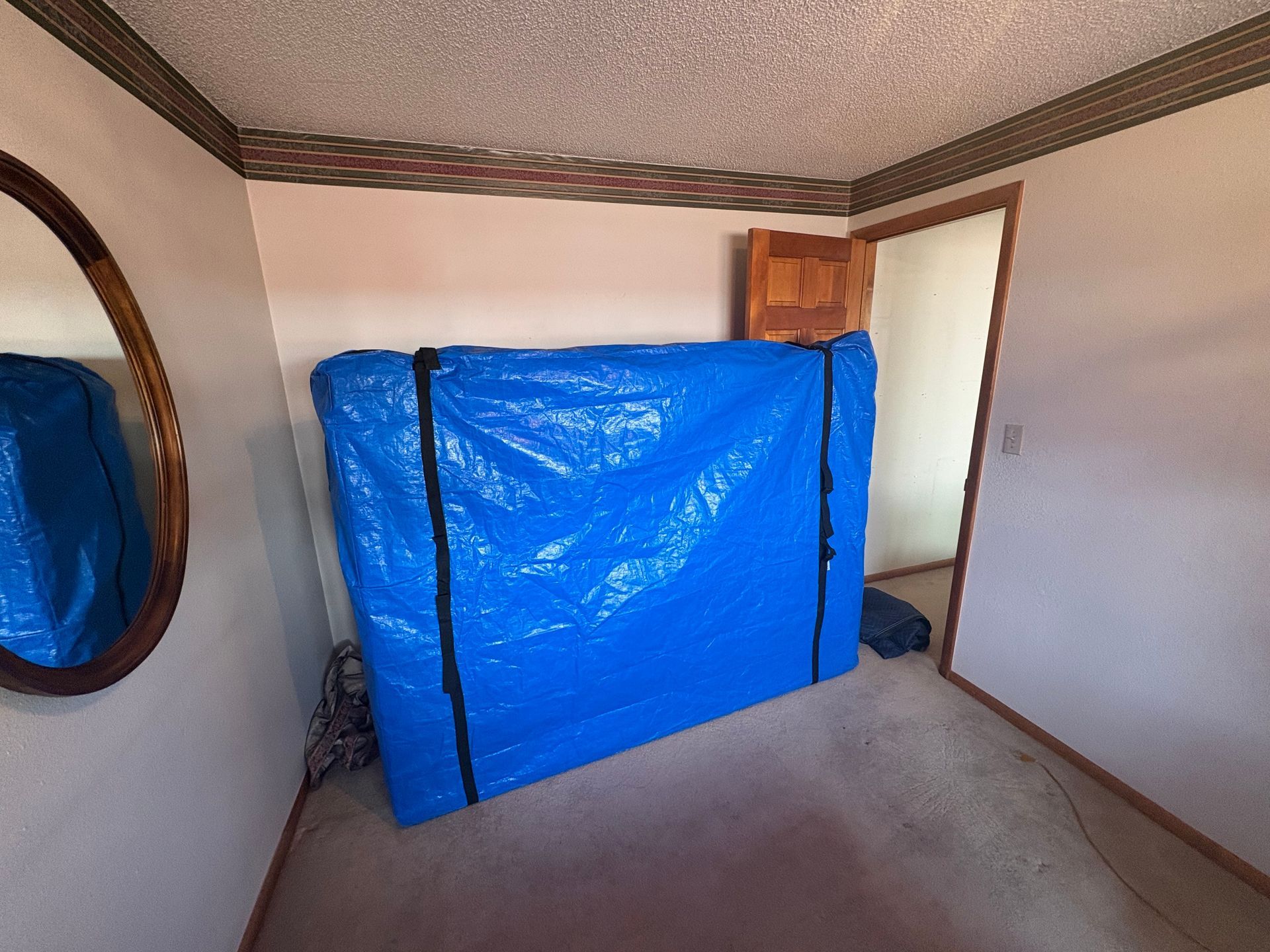 A large blue tarp covers an object, likely a mattress, in a room. Brown door, oval mirror.