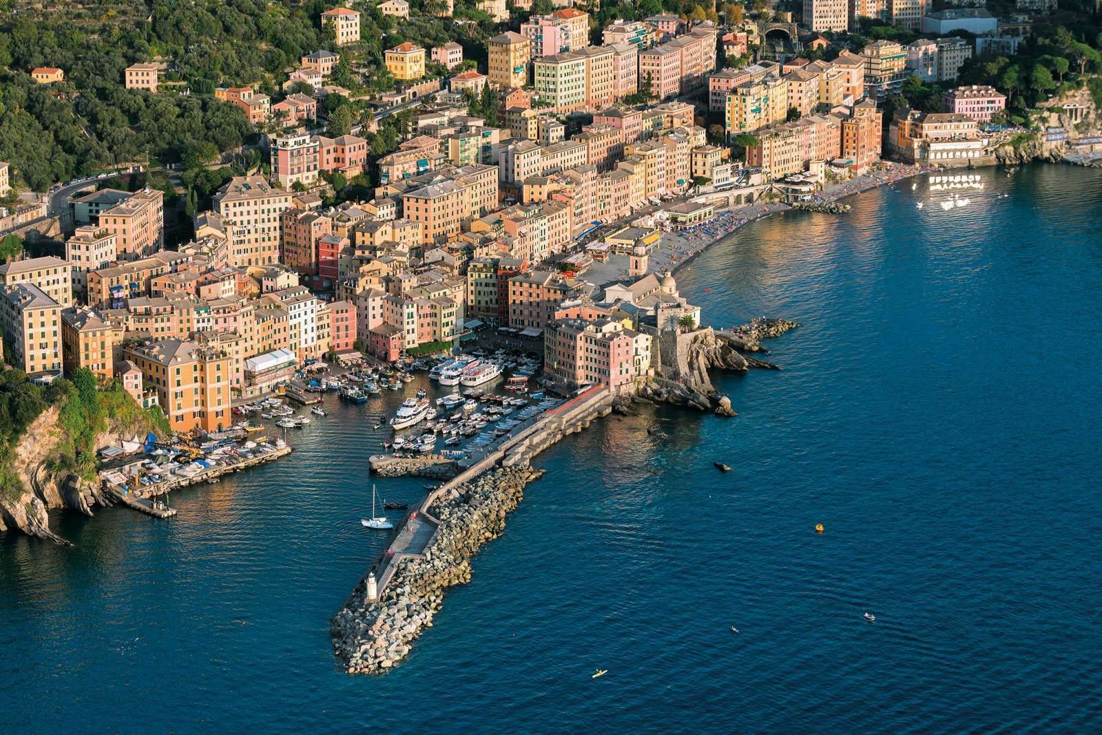 Città costiera con edifici colorati, porto con barche e frangiflutti rocciosi che si estendono nel mare blu.