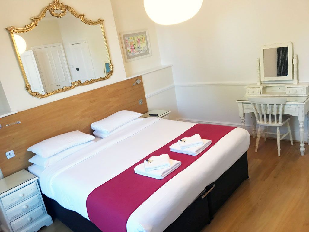 Bedroom with king-sized bed, decorative mirror, and vanity. White walls, wooden floor, and a pop of magenta on the bed.