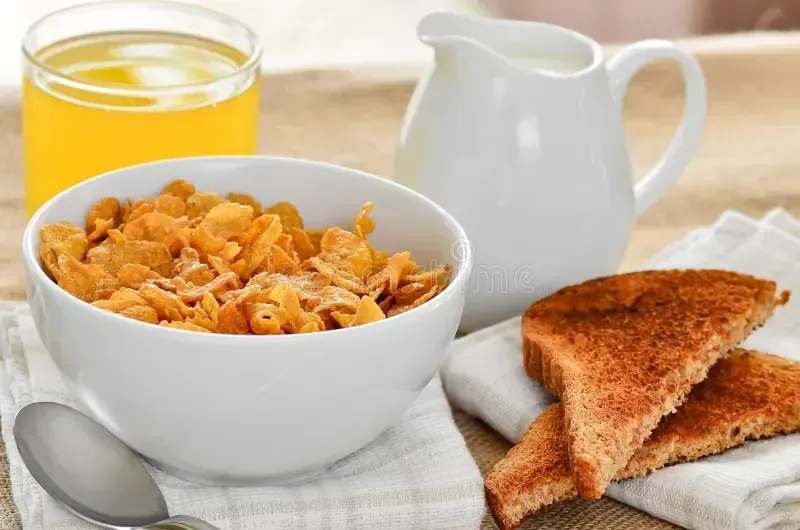 Bowl of cereal, toast, juice, and milk pitcher on a table.