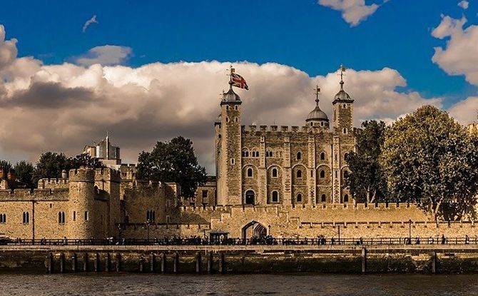 Tower of London, a stone fortress with turrets and a flag, against a cloudy blue sky, near water.
