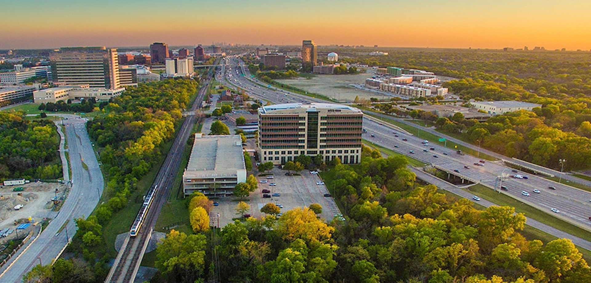 Aerial view of a city with buildings and roads surrounded by lush green trees under a colorful sunset.