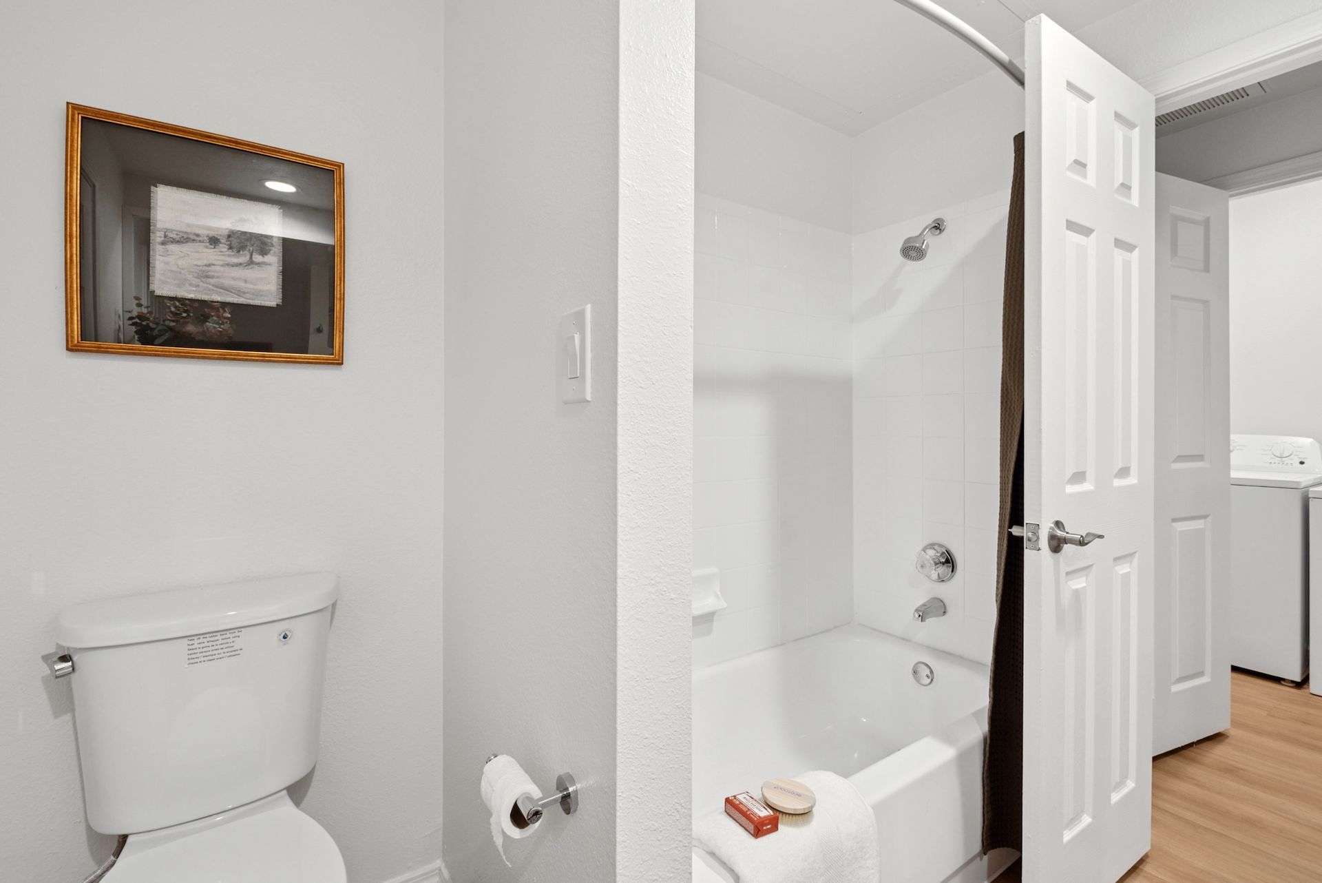 Bathroom with a white toilet, bathtub, and door; a picture hangs on the wall.