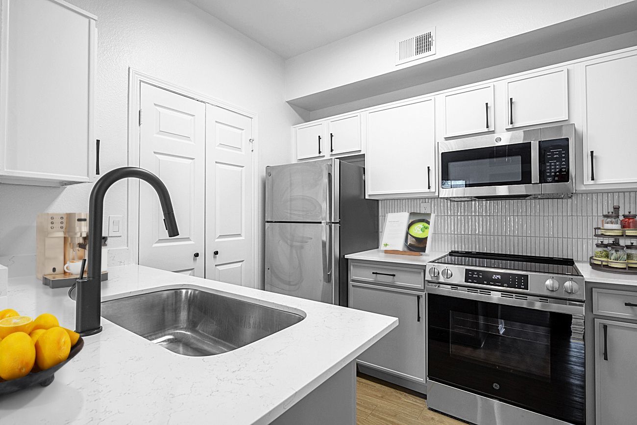 Modern kitchen with stainless steel appliances, white cabinets, and a gray countertop. A black faucet and lemons are visible.