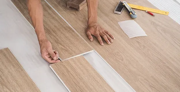 Hands installing light wood-look flooring. Tools and cut pieces visible. Vinyl Flooring Auckland
