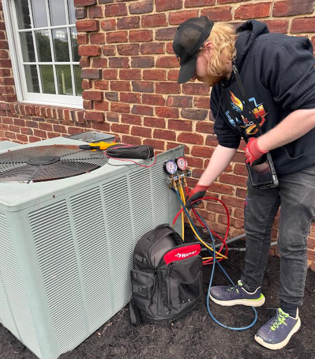 Person servicing HVAC unit outdoors with gauges and a tool bag beside a brick wall
