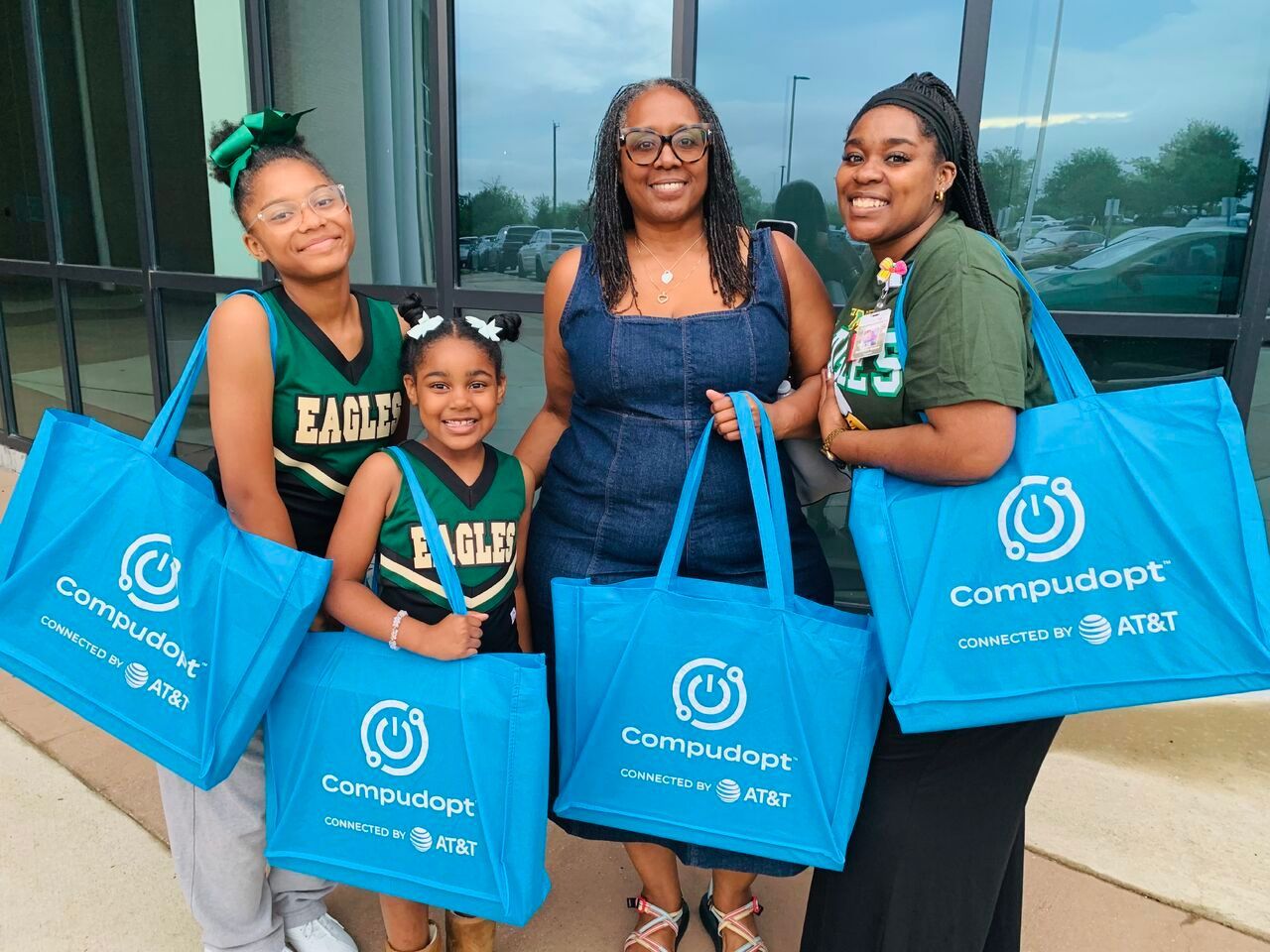 Four smiling people holding blue Compudopt bags outside a glass building