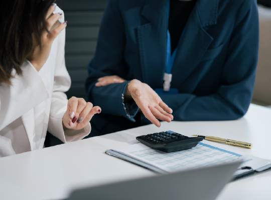 Two women are sitting at a table with a laptop and a calculator.