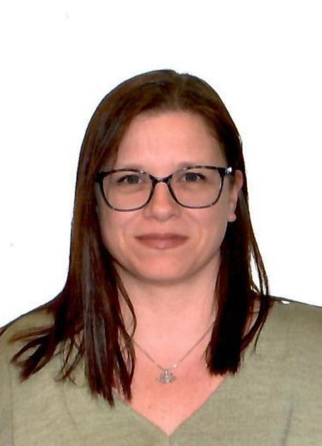 A person with shoulder-length brown hair and glasses, wearing a green V-neck shirt and a simple necklace.