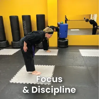 People in black martial arts uniforms perform a formal bow in a studio with yellow walls and punching bags.