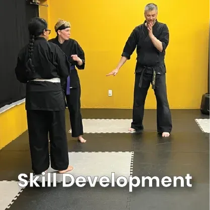 An instructor in a black gi demonstrates techniques to two students in a martial arts studio with yellow walls.