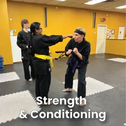 Two martial arts students in black uniforms practice sparring in a yellow-walled gym, observed by an instructor.