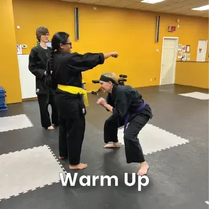 Two people practicing martial arts in a studio. One person, wearing a yellow belt, reaches out while another crouches down.
