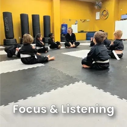 A class of children in martial arts uniforms sitting on a gym mat in a circle, listening to their instructor.