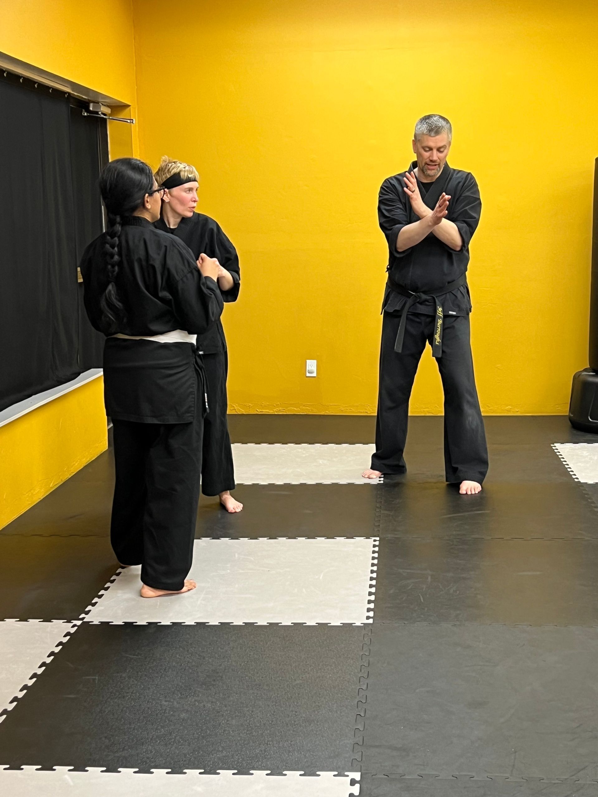 Three people wearing black martial arts uniforms practice a stance in a room with yellow walls and black-and-white matting.