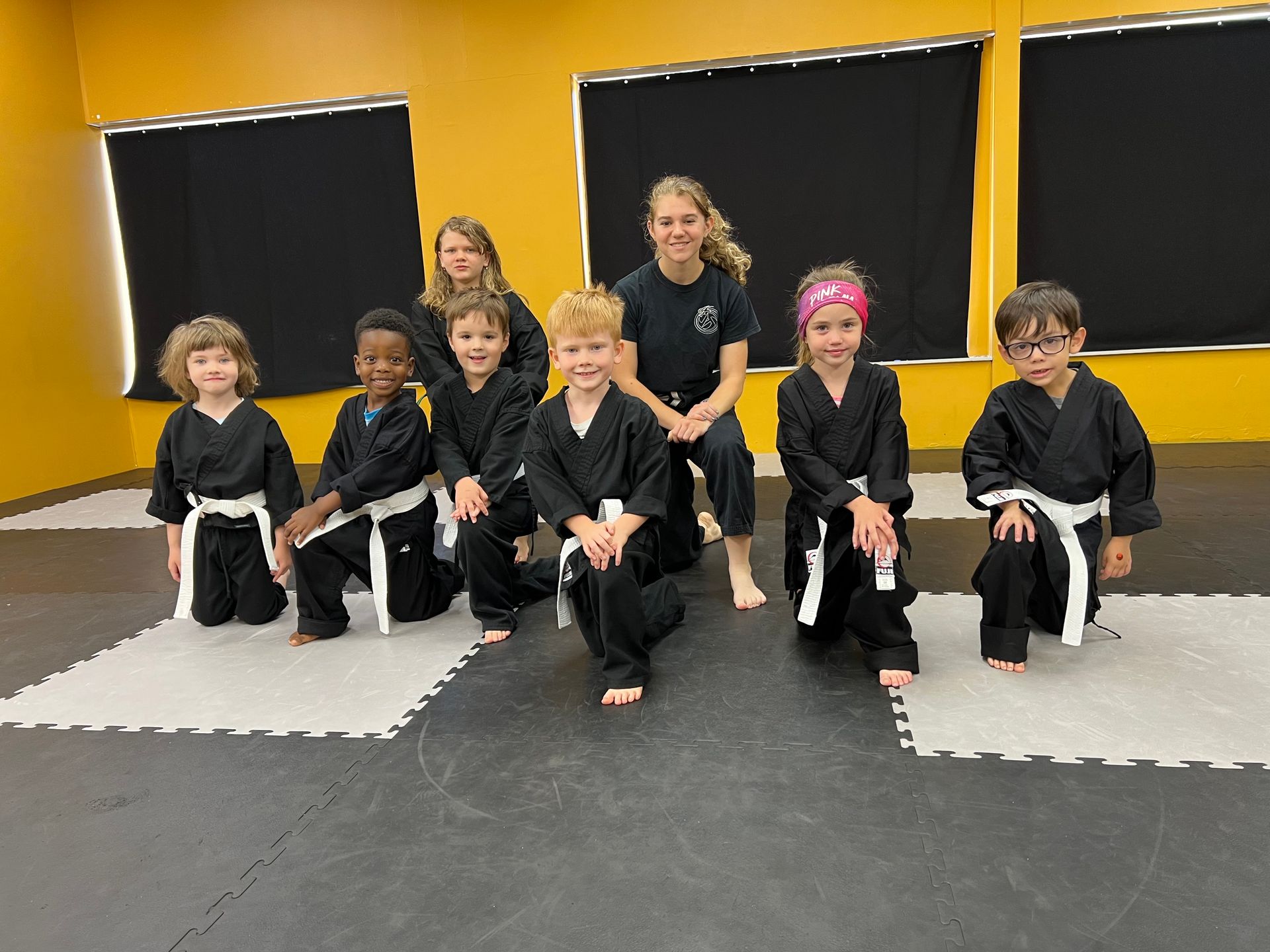 A group of children and an instructor in black martial arts uniforms pose on mats in a room with yellow walls.