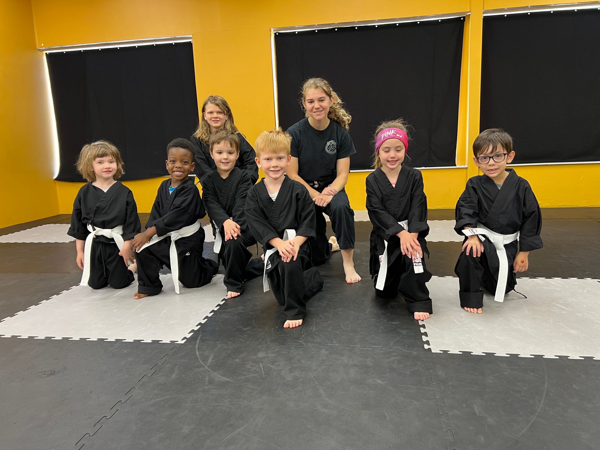 A group of students and their instructor in black karate uniforms kneel on gym mats in front of yellow walls.