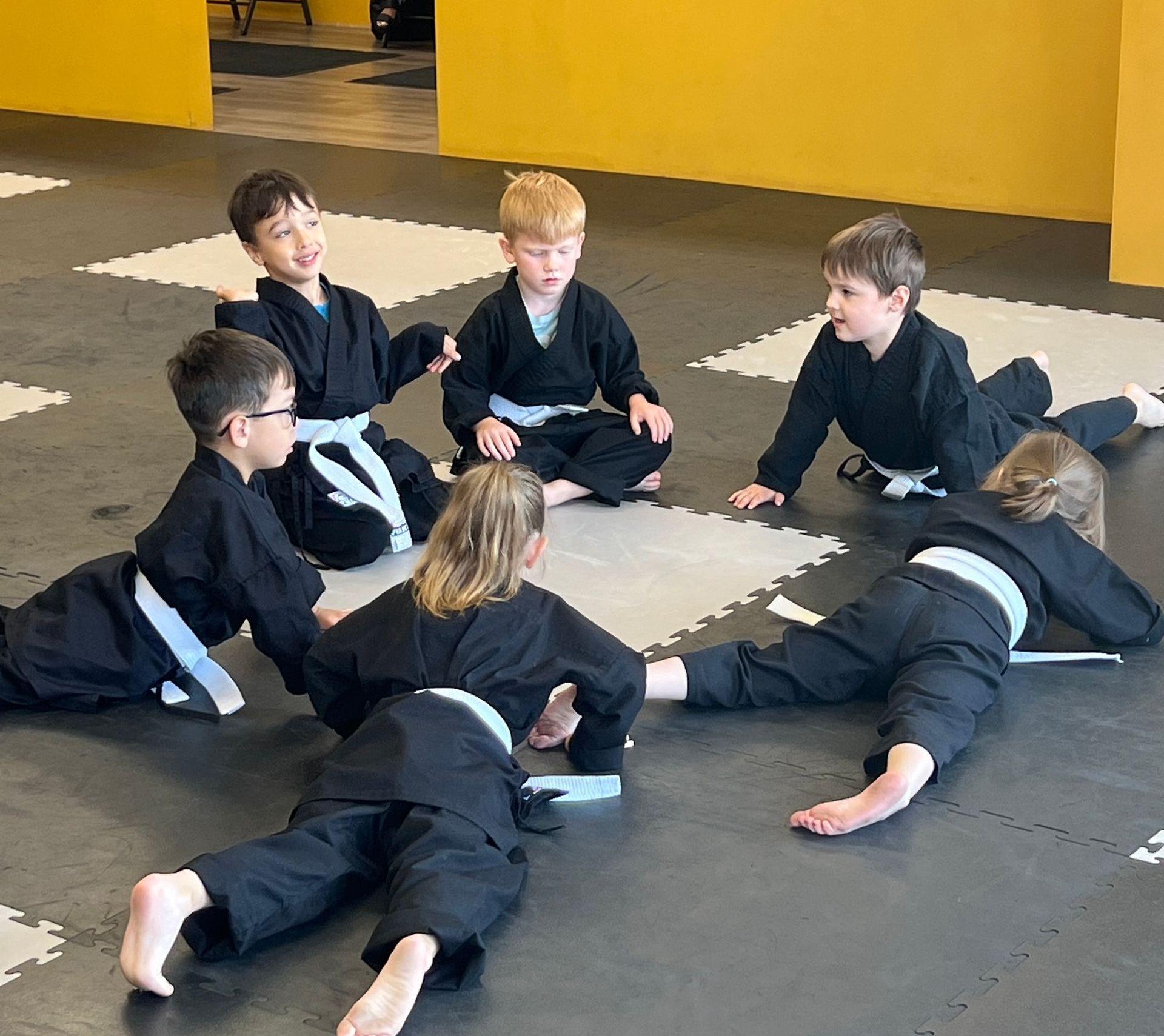 Six children in black martial arts uniforms with white belts sit or lie on gym mats during a class.