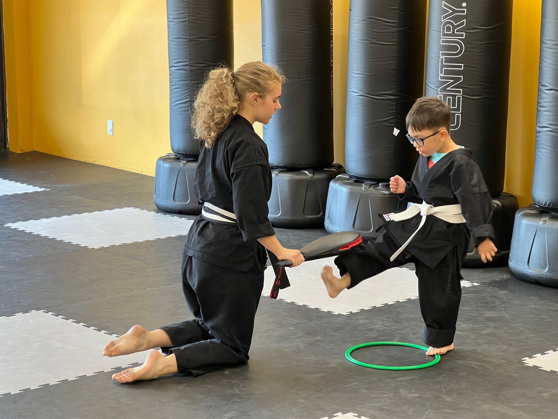 A martial arts instructor kneels while holding a target pad for a student practicing a kick inside a gym.