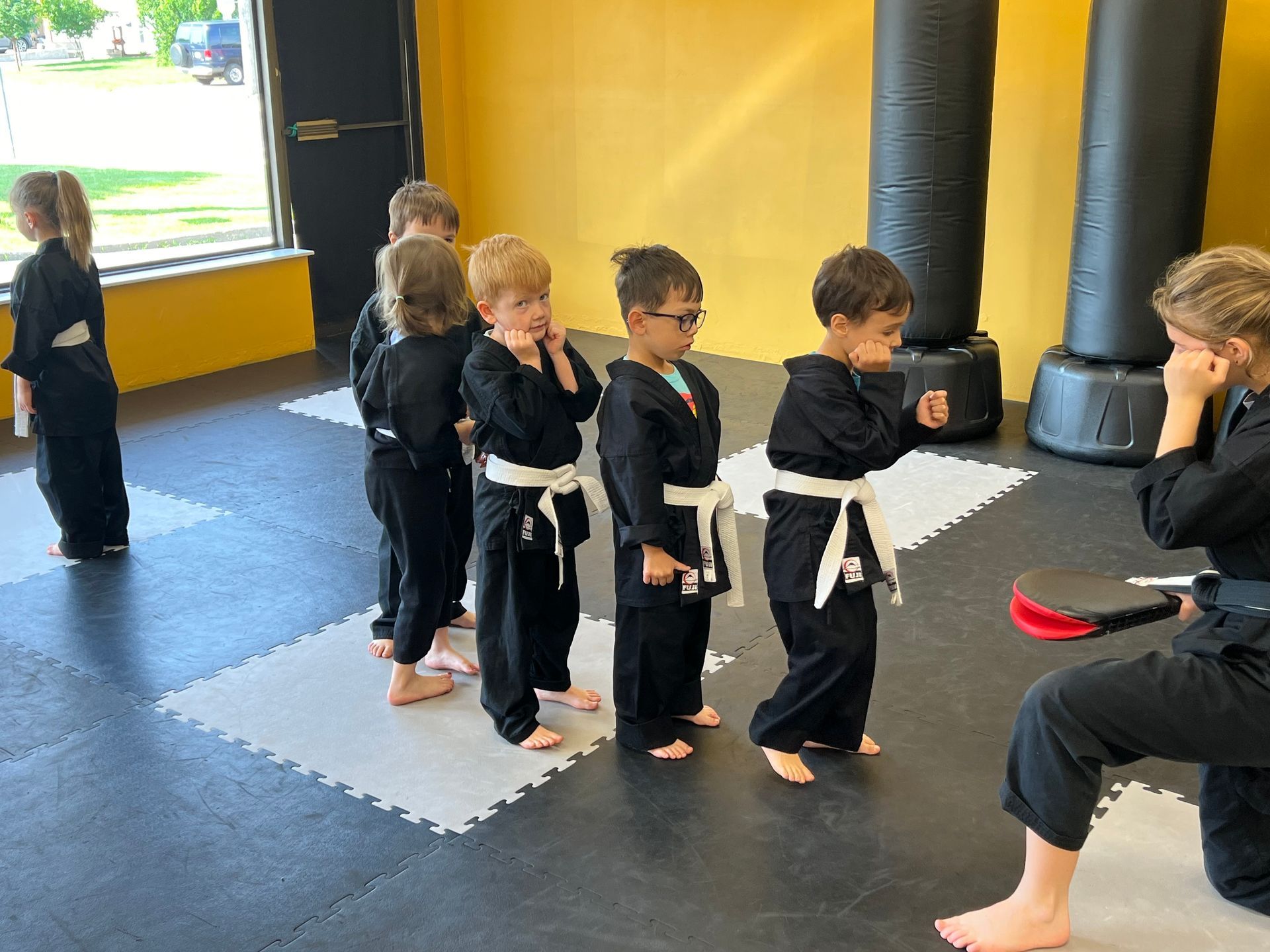 Children in black martial arts uniforms line up on floor mats in a studio while an instructor kneels with a practice pad.