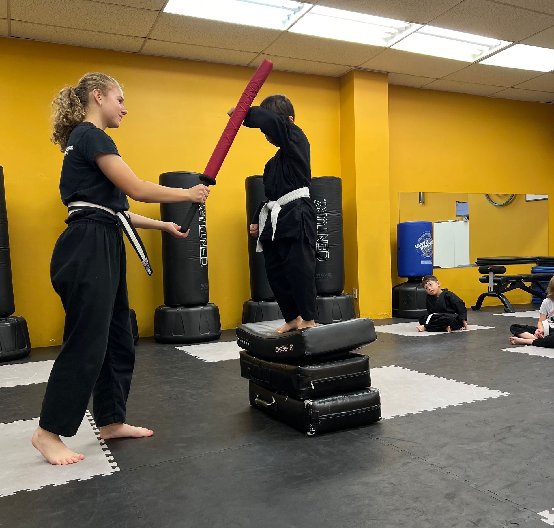 A martial arts instructor holding a foam practice sword while a student stands on a stack of training pads in a studio.