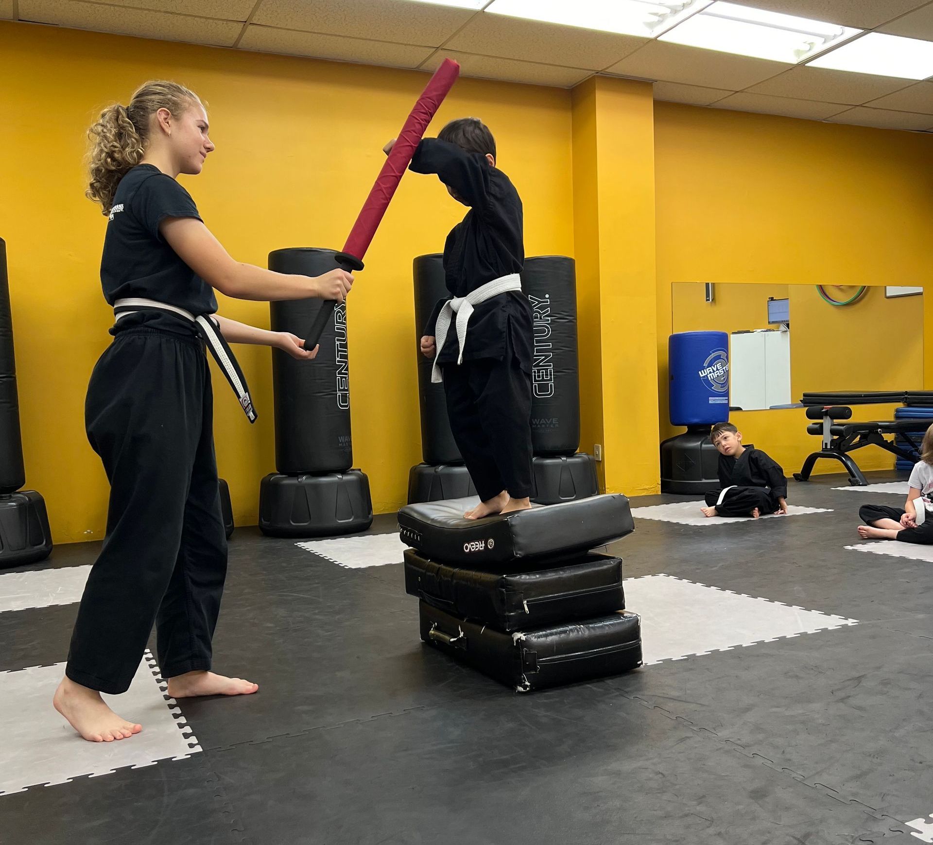 A martial arts instructor holds a practice sword over a student standing on stacked training pads in a gym.