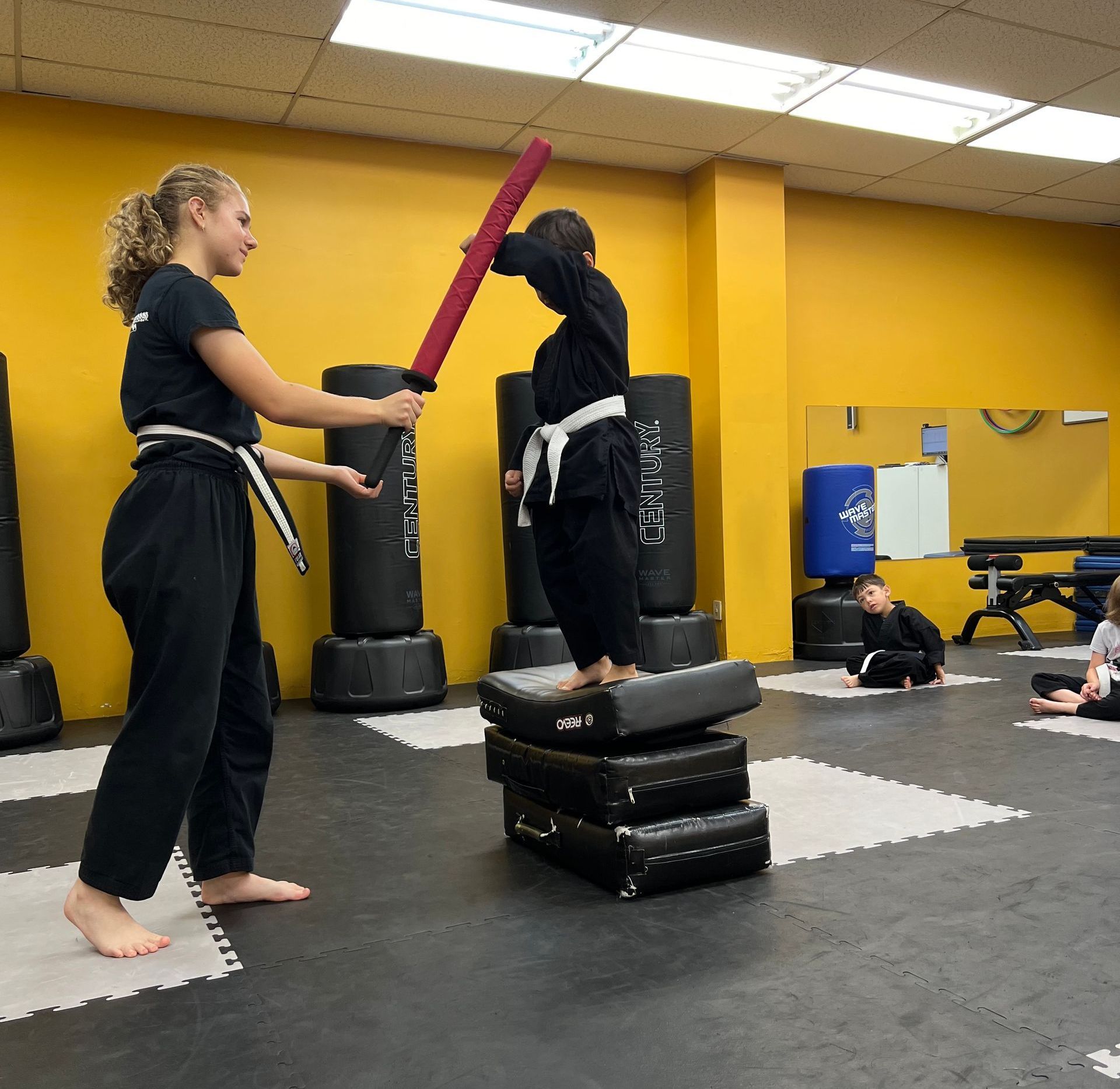 A martial arts instructor holding a foam training sword for a student standing on a stack of training mats in a studio.