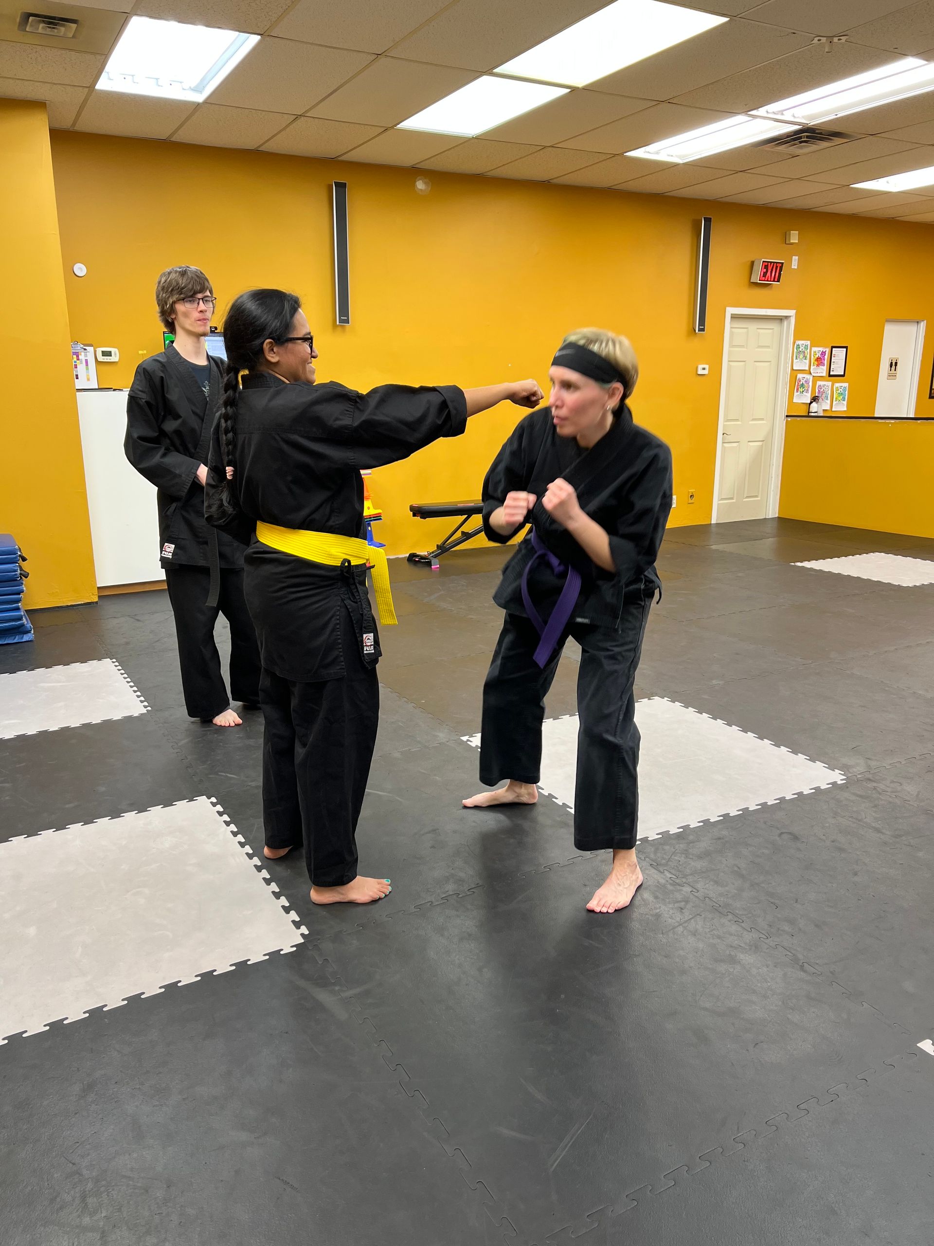 Two martial arts students in black uniforms practice a punch and block in a studio, watched by a third student.