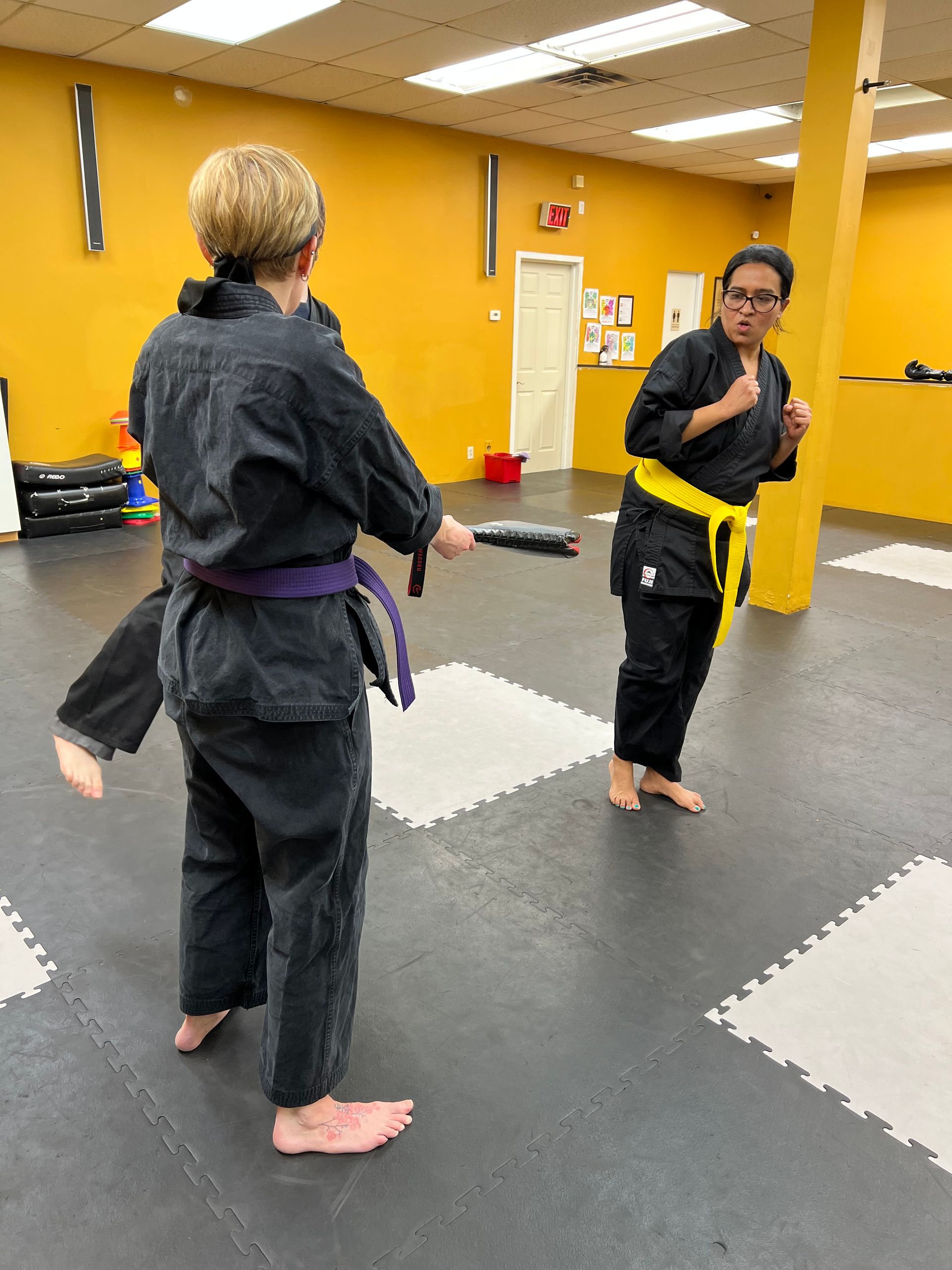 A person with a purple belt holds a target for a person with a yellow belt in a martial arts studio with yellow walls.