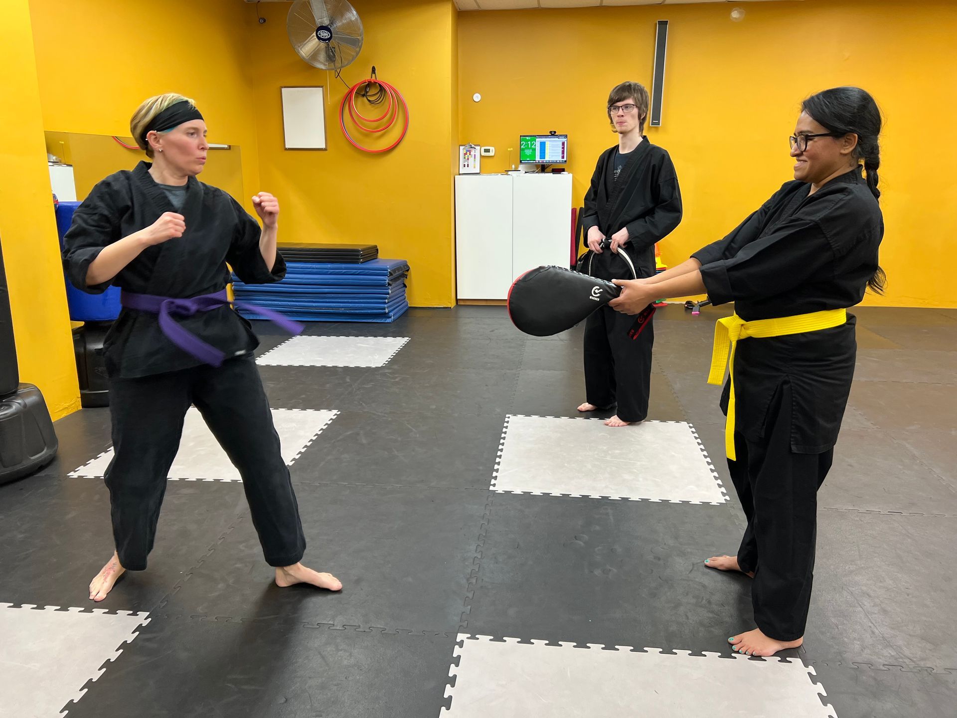 Three martial arts students in uniforms practice with a focus pad in a studio with yellow walls and puzzle-mat flooring.