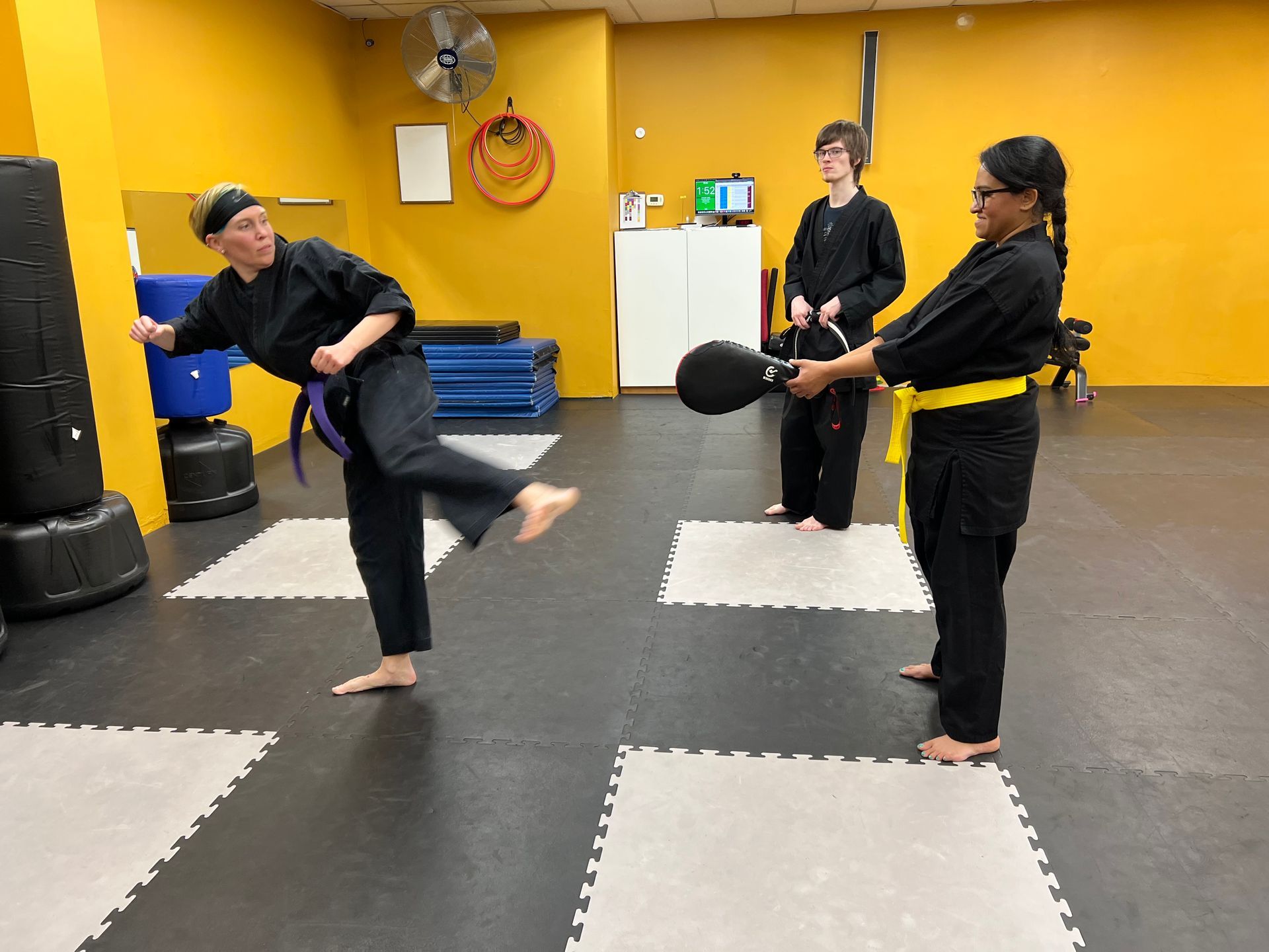 Two martial arts students in black uniforms practice kicking a training pad held by another student in a yellow-walled gym.