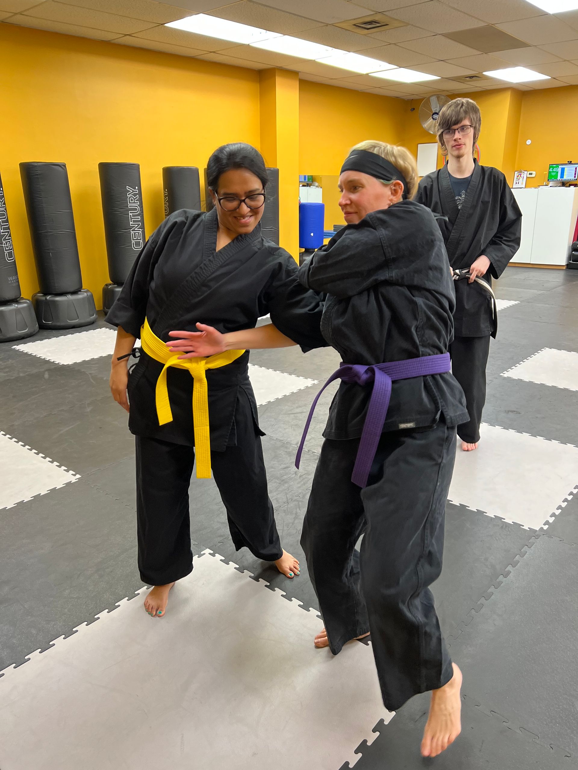 Two people in black martial arts uniforms practice self-defense moves in a gym, with an observer standing in the back.