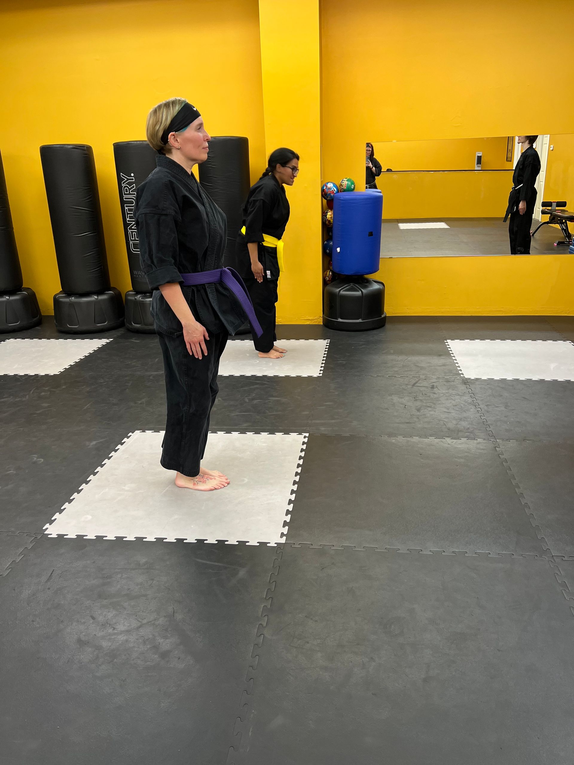 Two people in black martial arts uniforms and belts perform a standing bow inside a studio with yellow walls.