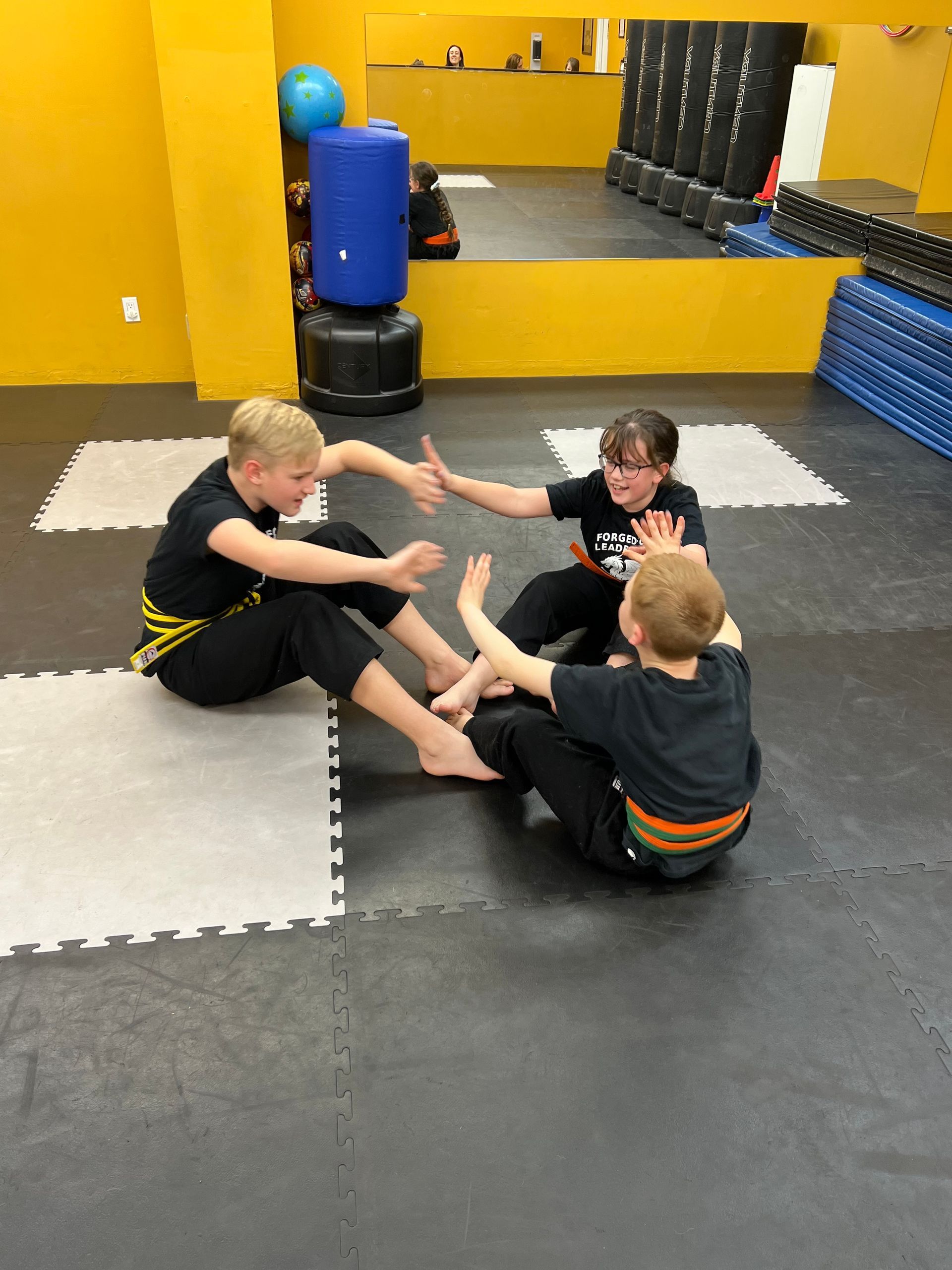 Three people in martial arts uniforms sit on a gym floor, reaching out to high-five each other in a circle.