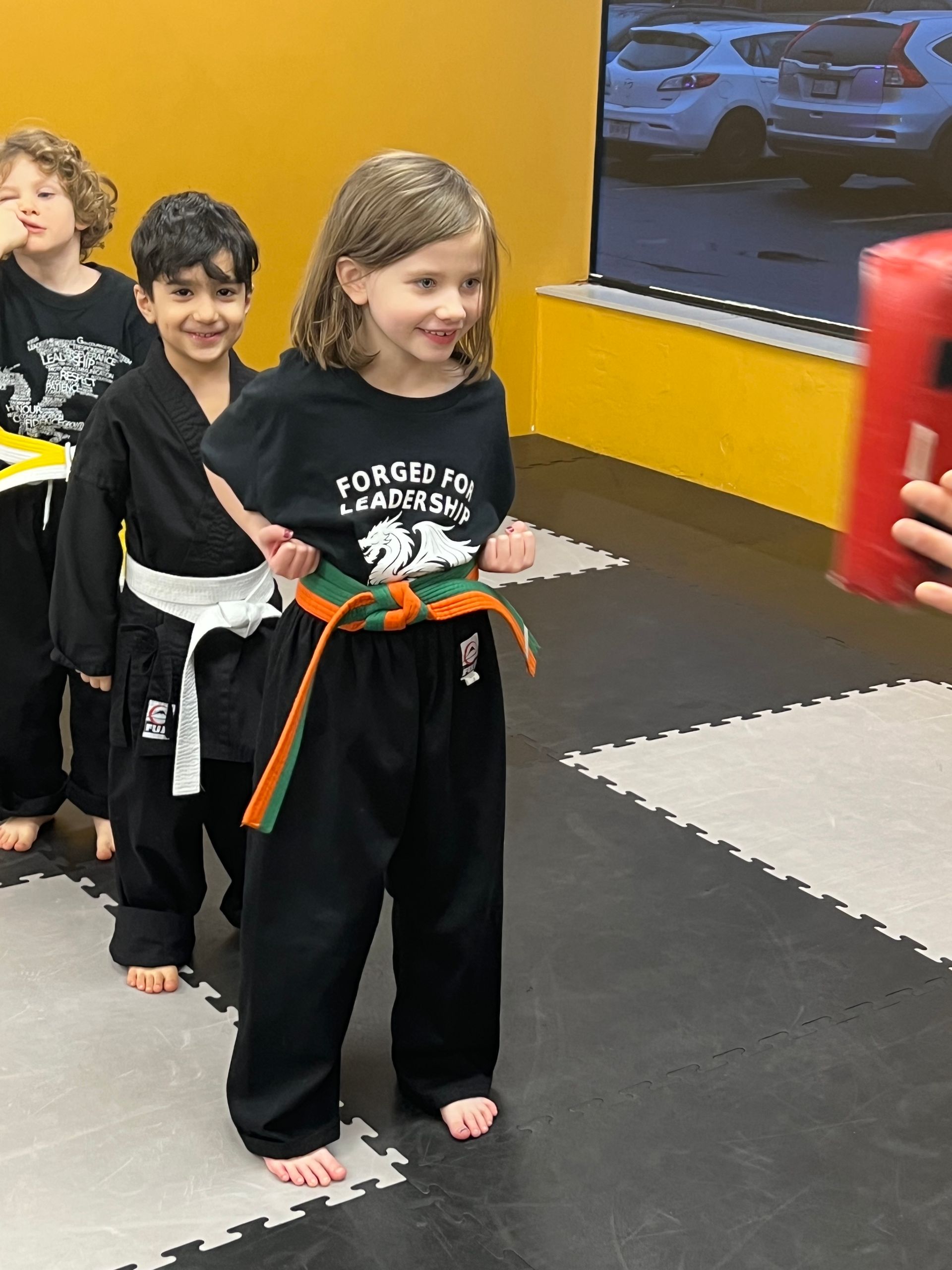 Three students in black martial arts uniforms practice in a studio with a red striking pad held in the foreground.