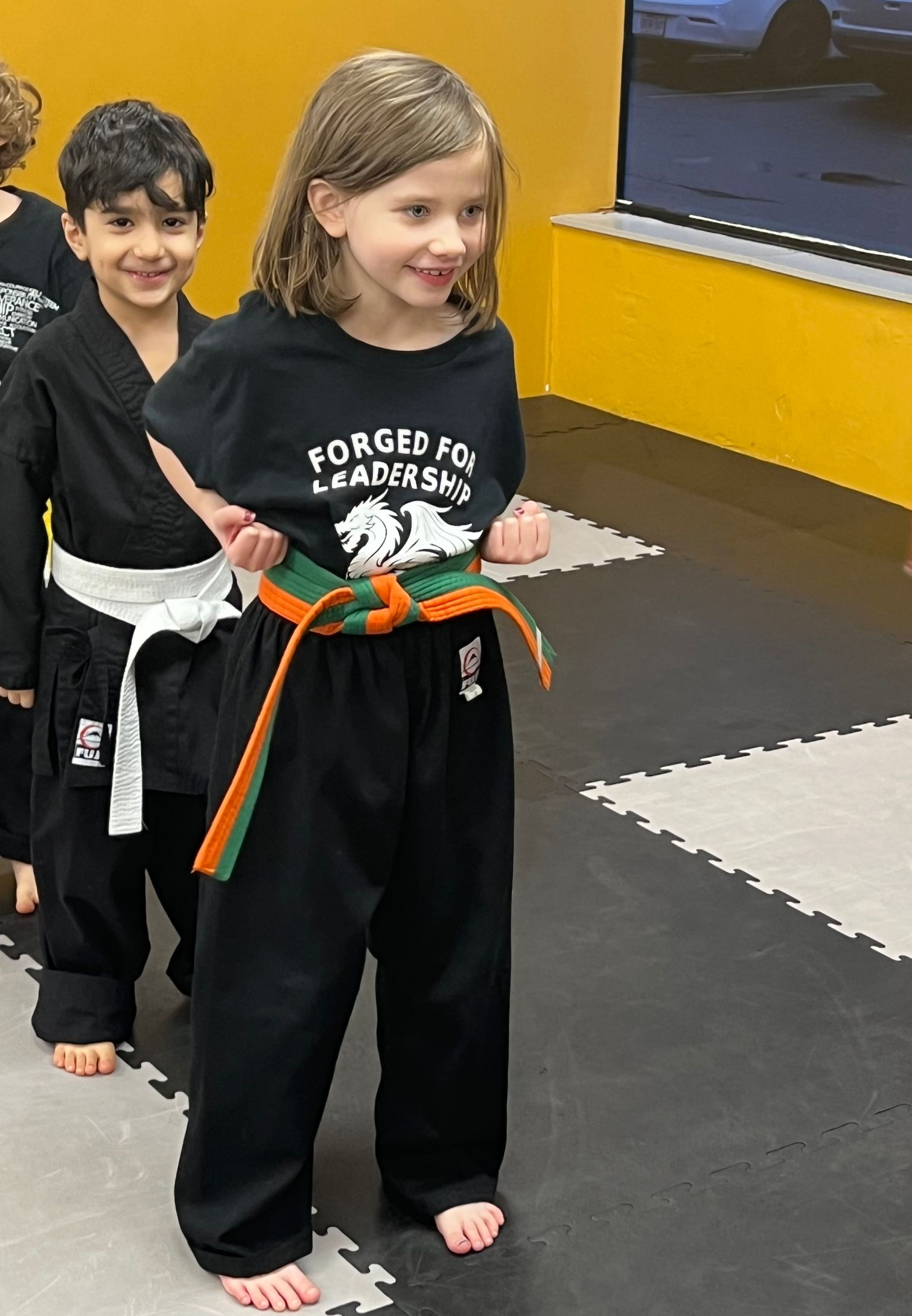 Two students in martial arts uniforms with colorful belts stand side-by-side in a practice studio.