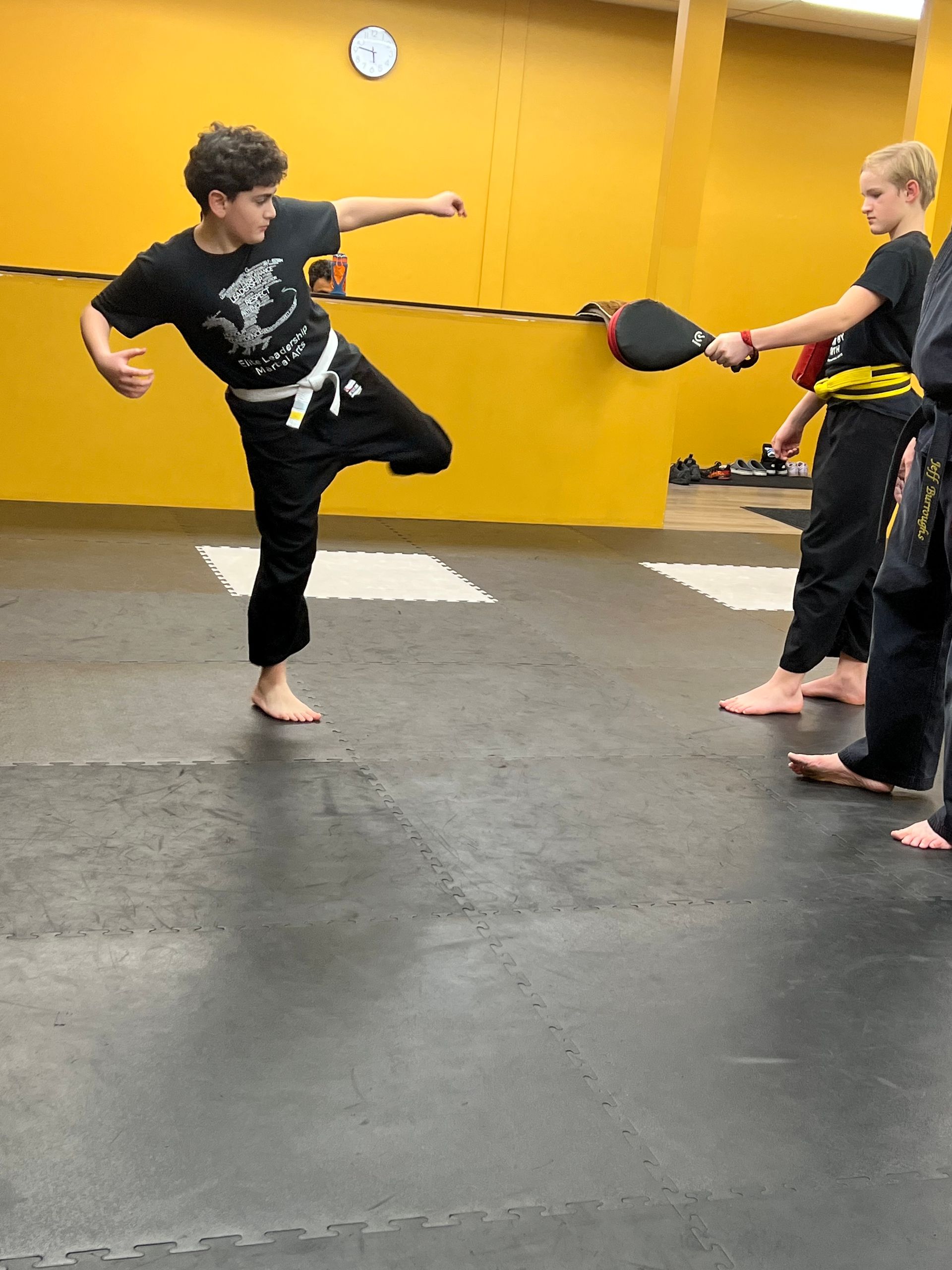 A student performs a side kick toward a target held by another student in a martial arts studio with yellow walls.