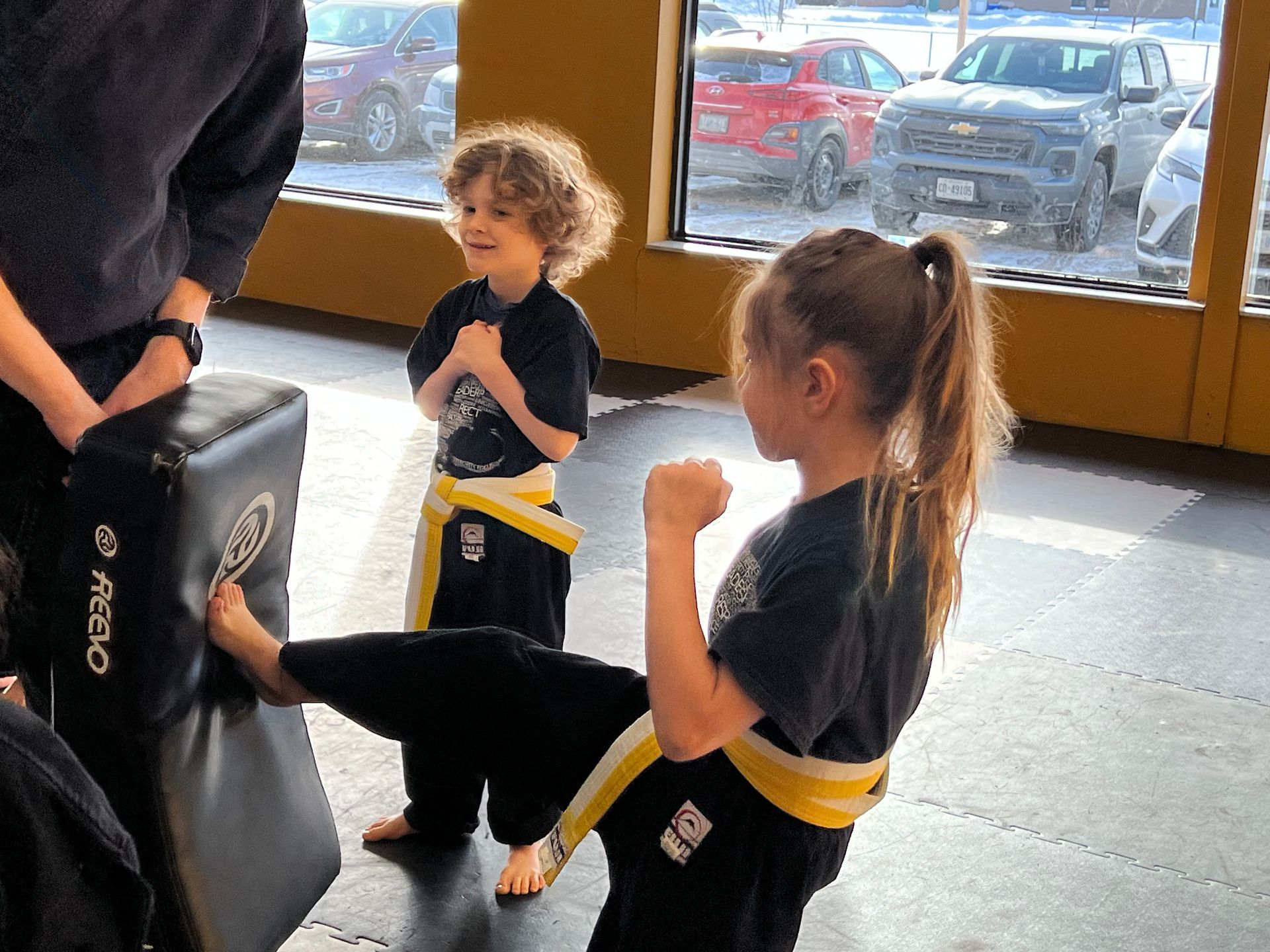 A group of children and an instructor in black martial arts uniforms pose on mats in a room with yellow walls.