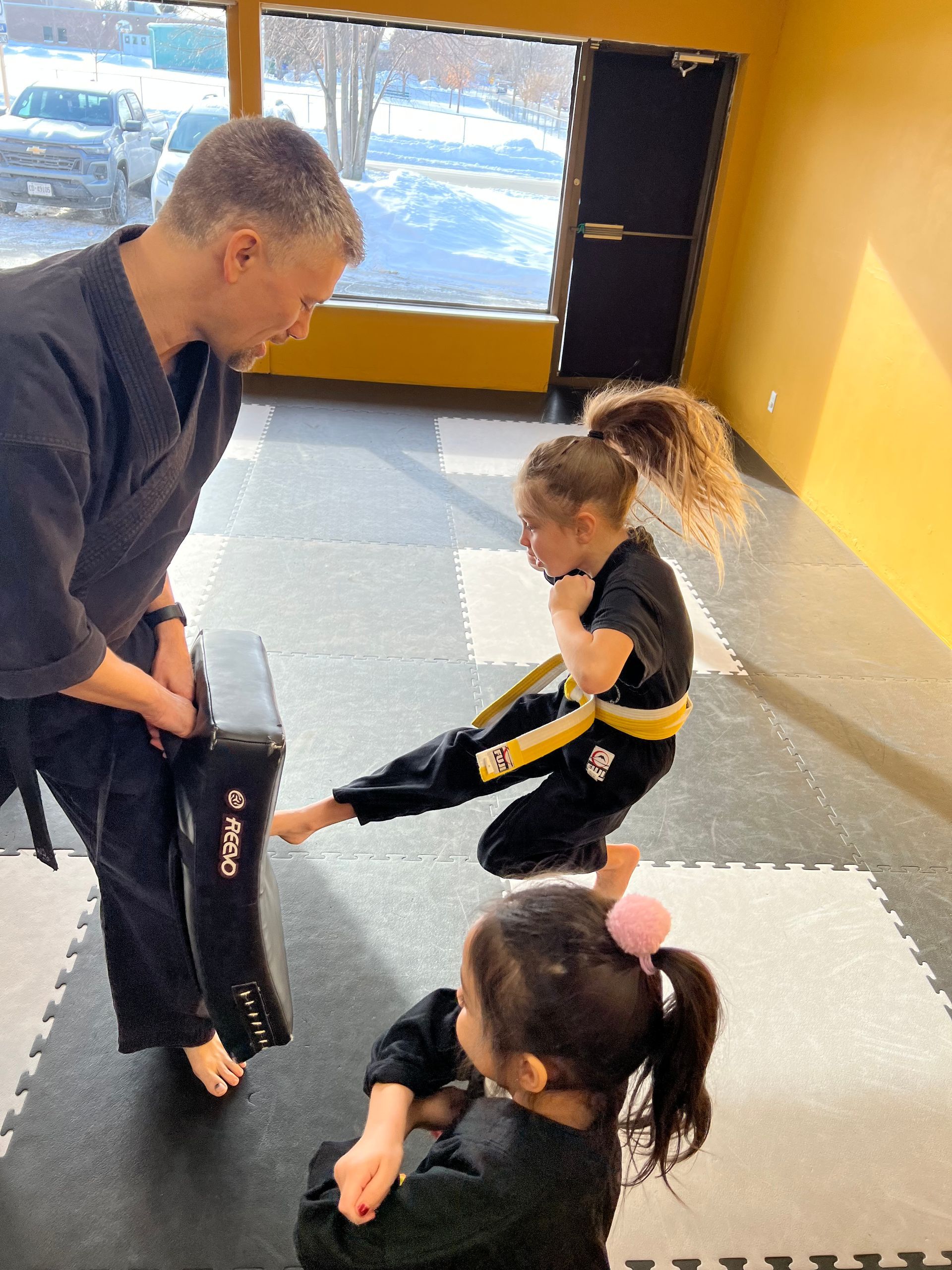 An instructor holds a black kicking pad for a student wearing a yellow belt, while another student watches in a studio.