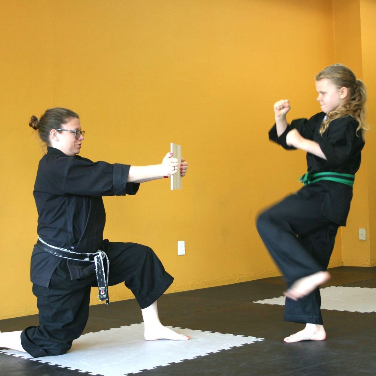 A person in a black uniform kneels holding a wooden board as a student prepares to kick it in a martial arts studio.