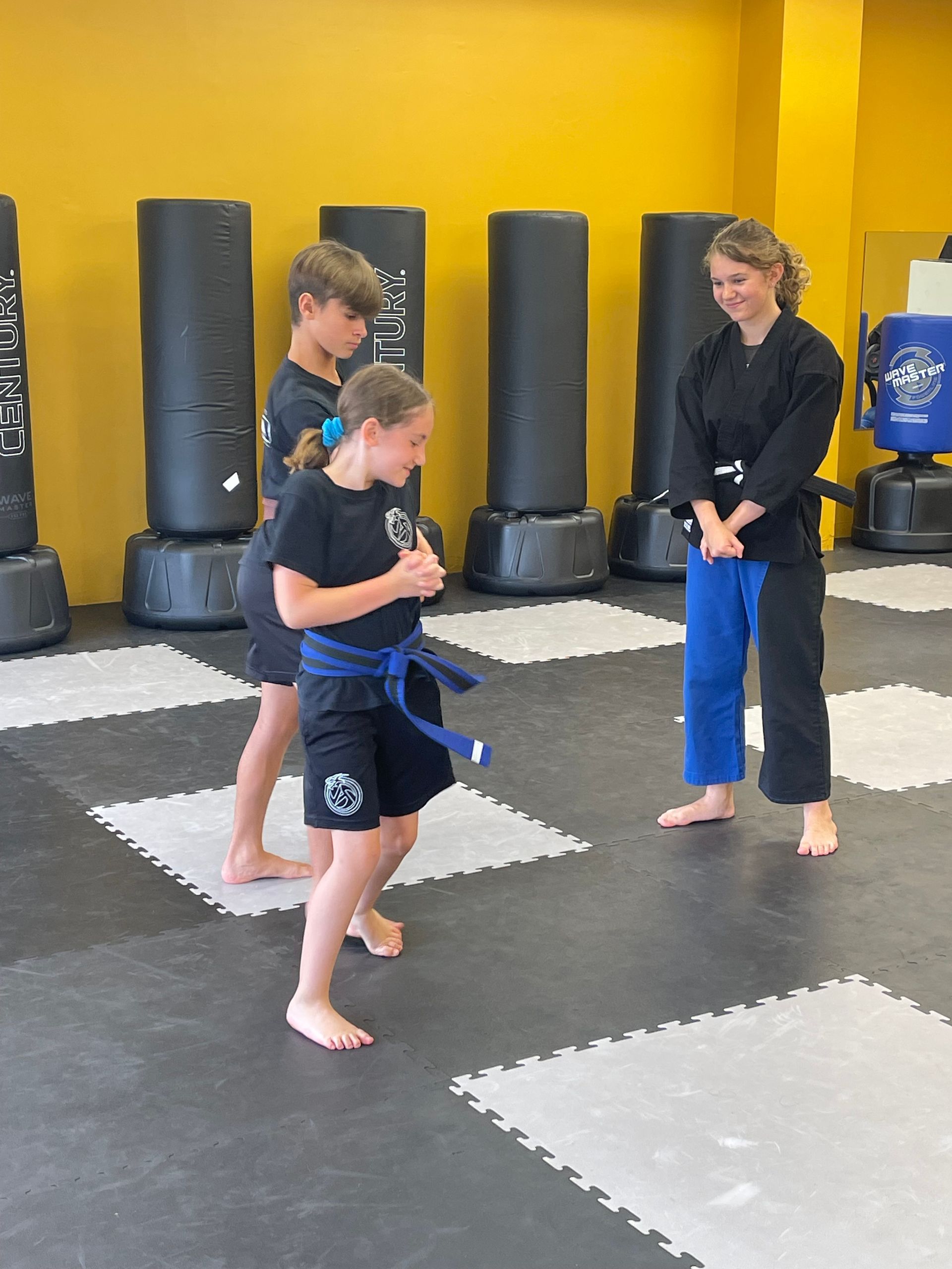 Two students in martial arts uniforms practice on a gym floor with a training partner watching nearby.