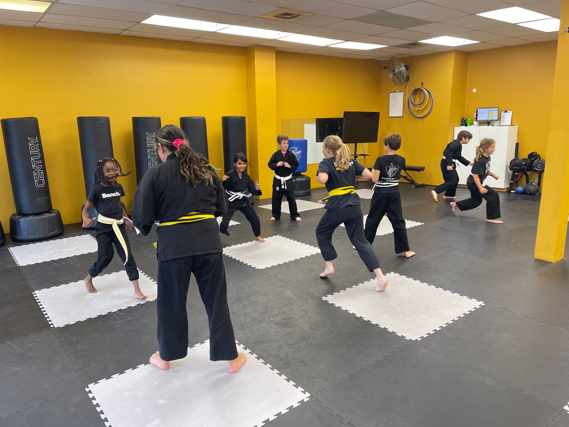 A group of students wearing martial arts uniforms and yellow belts practice stances in a studio with foam floor tiles.