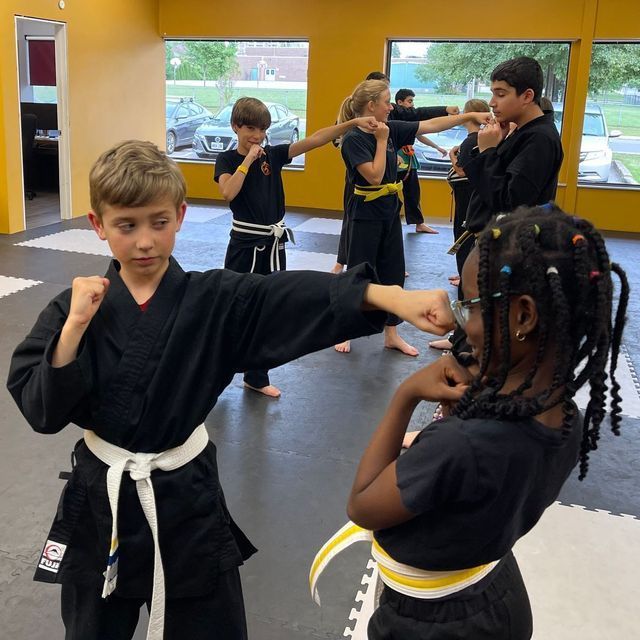 Children in martial arts uniforms practice punches in a gym with yellow walls and large windows.