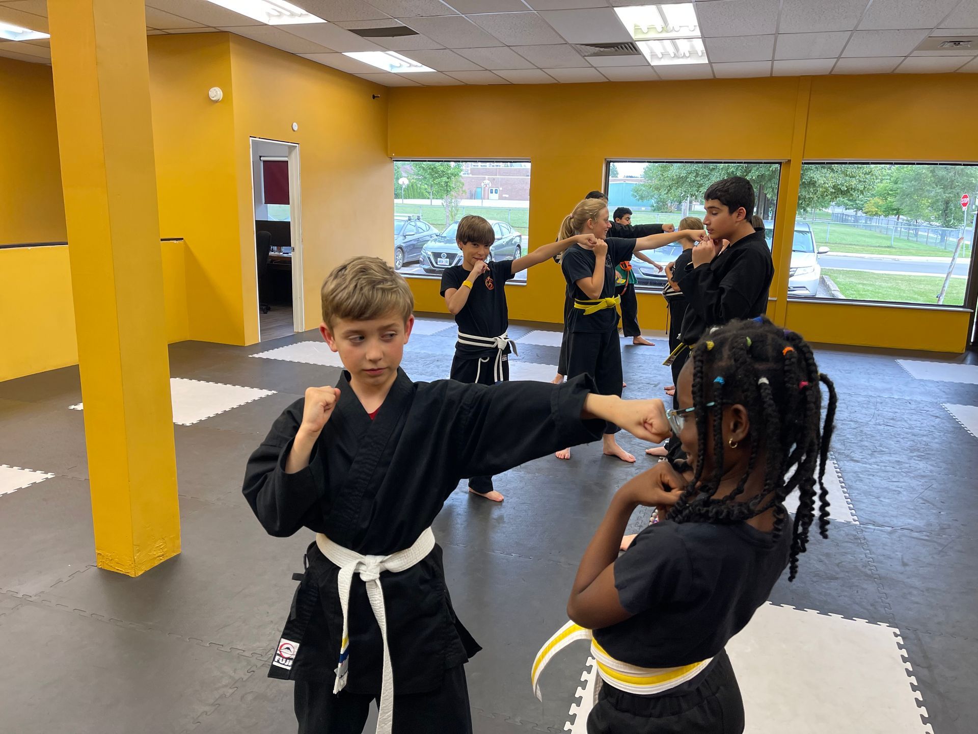 Martial arts students in black uniforms practice sparring techniques inside a yellow-walled training studio.