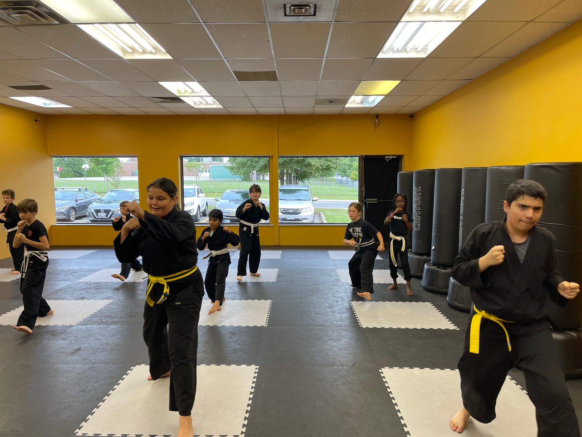 Students in black uniforms with yellow belts practice martial arts poses in a gym with square floor mats and tall bags.
