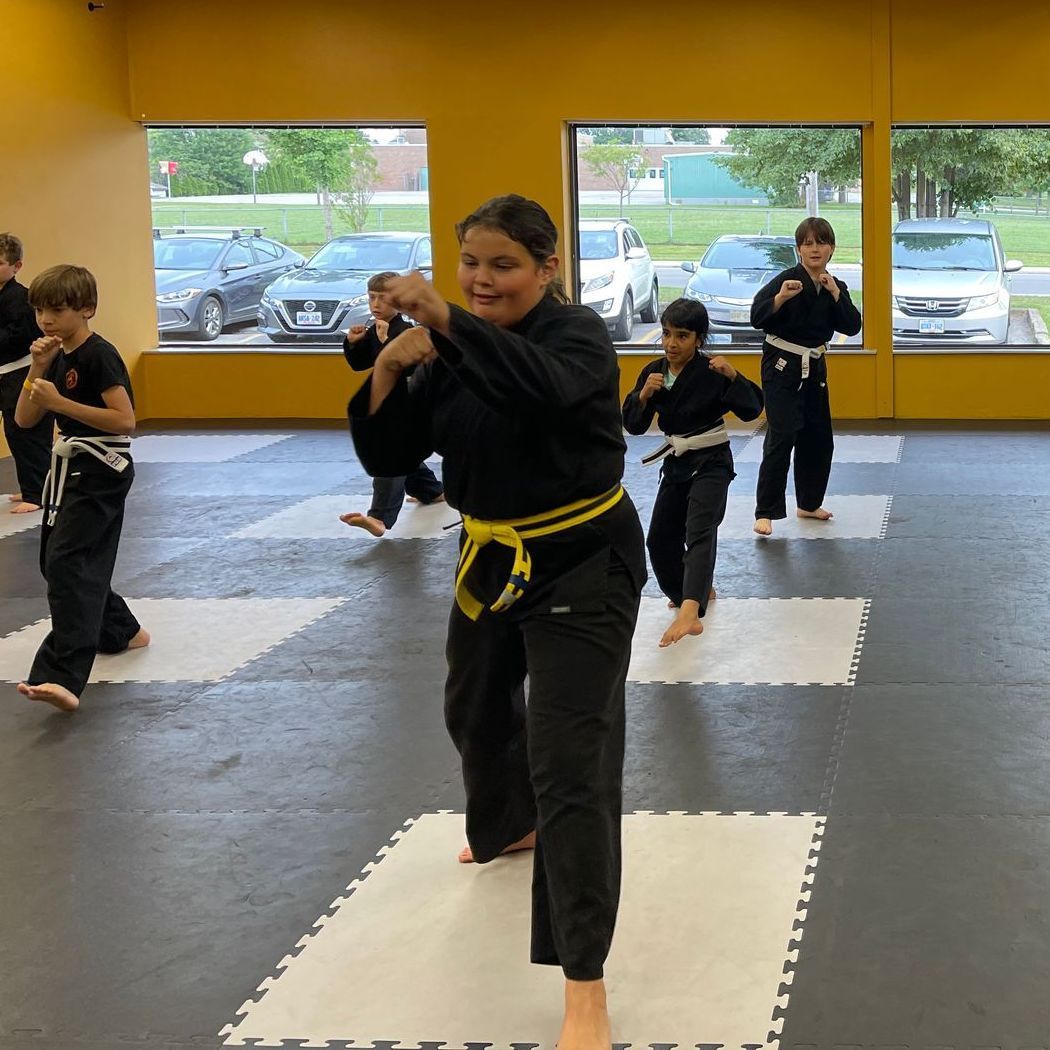 Children in black martial arts uniforms with colored belts practice defensive stances on floor mats in a training studio.