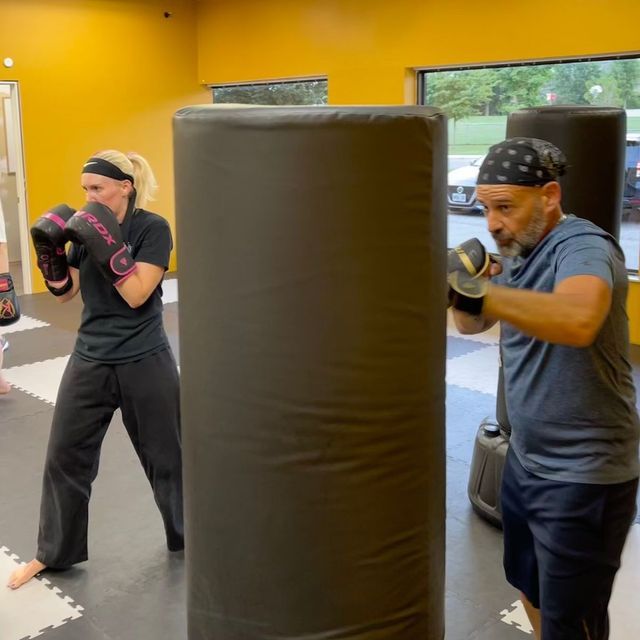 Two people in a gym practice boxing on tall black punching bags while wearing gloves and headbands.