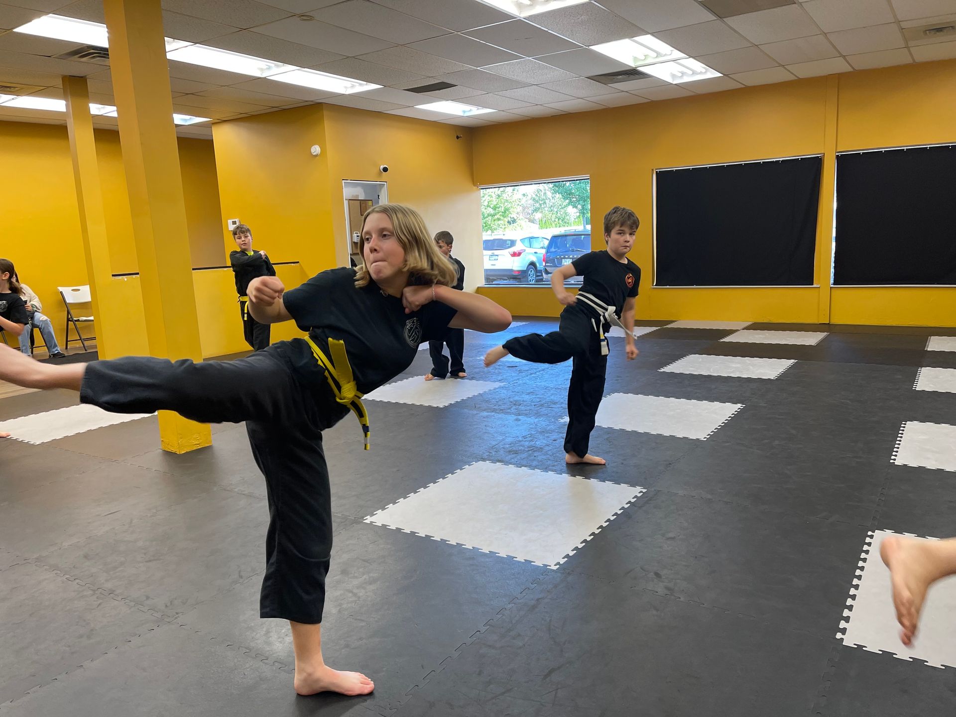Students in black uniforms perform side kicks in a bright training studio with yellow walls and black and white floor mats.