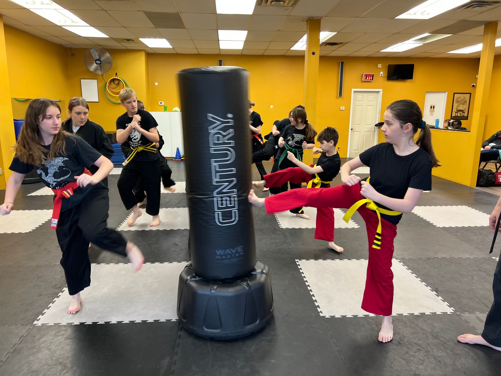 Students in black uniforms with yellow belts practice martial arts poses in a gym with square floor mats and tall bags.